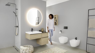 Un hombre camina en un baño con lavabo, WC, bidé y muebles de la serie Geberit iCon Un hombre camina en un baño con lavabo, WC, bidé y muebles de la serie Geberit iCon
