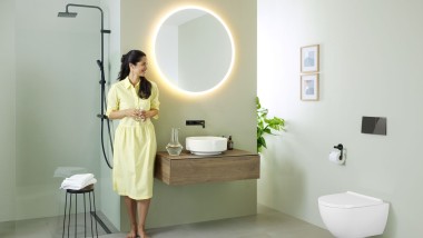 Mujer con vestido amarillo de pie delante de un baño verde menta con muebles y cerámica de baño de Geberit y grifería negra.